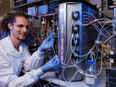 A man in a white coat and blue gloves sits in front of a silver cannister 